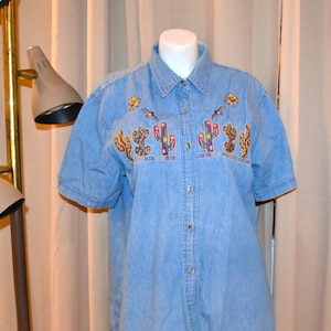 Puede incluir: Una camisa vaquera azul claro con mangas cortas y cierre de botones. La camisa presenta diseños de cactus bordados de colores y flores amarillas.