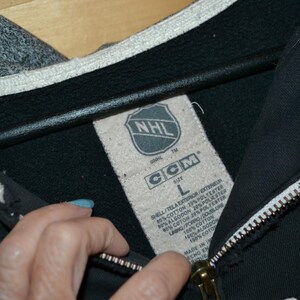 K&ouml;nnte beinhalten: Schwarzer Kapuzenpullover mit Rei&szlig;verschluss, grauer Kapuze und wei&szlig;em Besatz. Das Etikett zeigt das NHL-Logo und die Marke CCM. Der Rei&szlig;verschluss ist wei&szlig; mit einem goldfarbenen Zipper. Der Hoodie ist Gr&ouml;&szlig;e L.