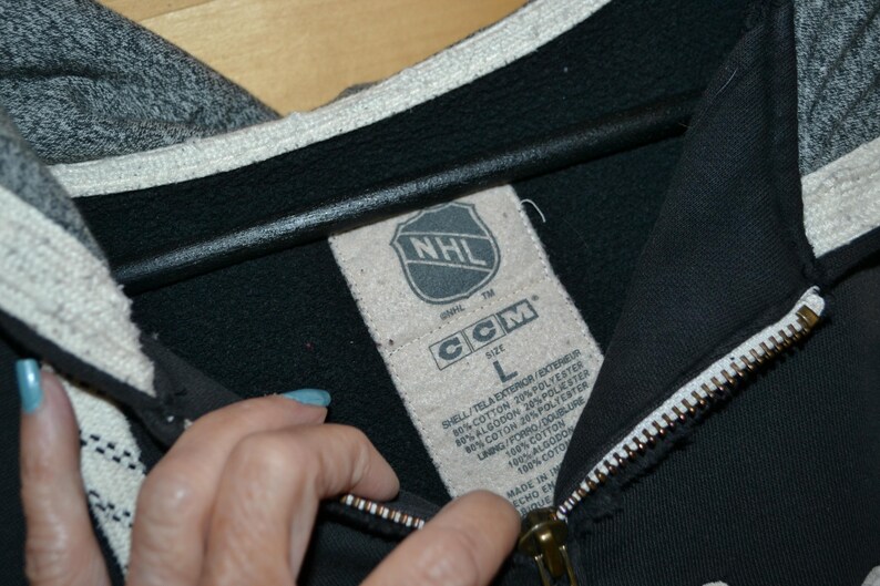 K&ouml;nnte beinhalten: Schwarzer Kapuzenpullover mit Rei&szlig;verschluss, grauer Kapuze und wei&szlig;em Besatz. Das Etikett zeigt das NHL-Logo und die Marke CCM. Der Rei&szlig;verschluss ist wei&szlig; mit einem goldfarbenen Zipper. Der Hoodie ist Gr&ouml;&szlig;e L.