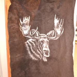 May include: A dark brown rectangular blanket featuring a large, detailed white graphic of a moose head with prominent antlers centered on the fabric. The blanket has a soft, textured surface and is suspended by three colorful plastic clips.