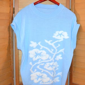 May include: A light blue sleeveless sweater with a white floral design. The sweater is hanging on a red hanger against a wooden background. The floral pattern is a repeating design of white flowers and stems.