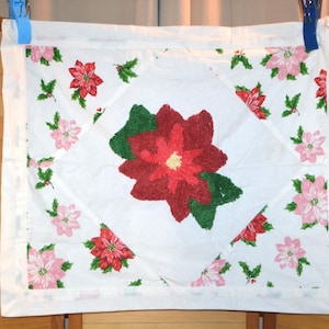 Pode incluir: Toalha de mesa quadrada branca com um desenho central de estrela-do-natal vermelha e verde. Os cantos e as bordas são adornados com estrelas-do-natal vermelhas e rosas menores e folhas verdes de azevinho. A toalha de mesa é presa por clipes azuis e brancos.