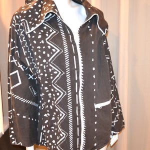 May include: A dark brown hooded jacket with a white geometric pattern. The jacket has a full zipper, white trim, and a drawstring at the bottom. The design includes dots, zigzags, and cross shapes.