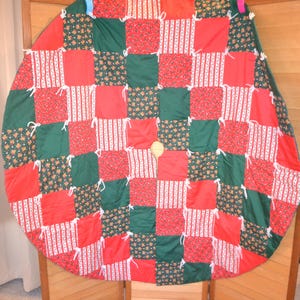 May include: A circular Christmas tree skirt with a patchwork design. It features red, green, and white fabric squares. Some squares have floral patterns, while others have stripes. White ties are sewn at the seams.