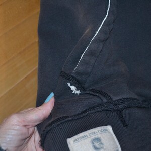K&ouml;nnte beinhalten: Nahaufnahme eines dunkelgrauen Kapuzenpullovers mit einer wei&szlig; gen&auml;hten Naht und einem kleinen gestickten Vogeldetail. Der Pullover hat unten ein Etikett mit Text.