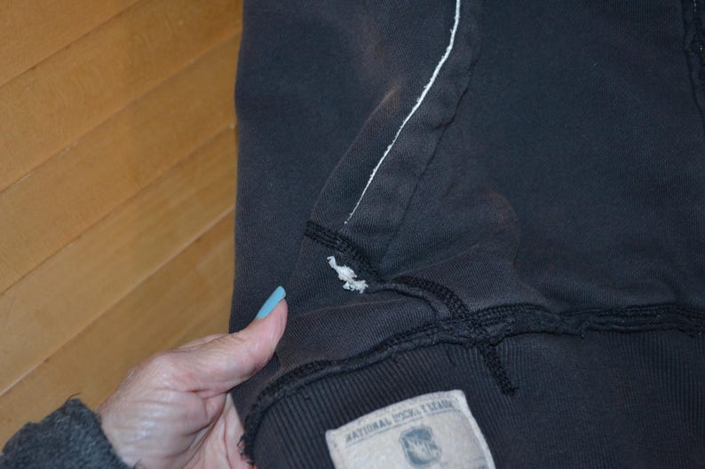 K&ouml;nnte beinhalten: Nahaufnahme eines dunkelgrauen Kapuzenpullovers mit einer wei&szlig; gen&auml;hten Naht und einem kleinen gestickten Vogeldetail. Der Pullover hat unten ein Etikett mit Text.