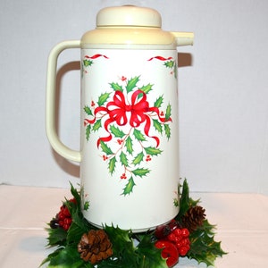 Può includere: Termos vintage color crema con un design a agrifoglio e nastro rosso. Il thermos ha un manico e un coperchio color crema. La base del thermos è circondata da una ghirlanda di agrifoglio con pigne e bacche rosse. Un oggetto festivo.