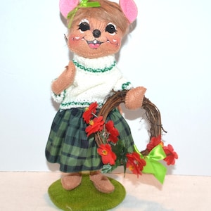 Könnte beinhalten: Eine skurrile Mäusefigur, die einen weißen Pullover mit grünen Verzierungen und einen grünen karierten Rock trägt. Die Maus hält einen Kranz mit roten Blumen und einer grünen Schleife. Die Figur steht auf einem grünen, kreisförmigen Sockel.