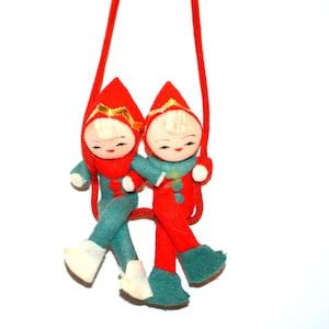Peut inclure: Deux ornements de Noël vintage en forme d'elfes. Chaque elfe porte un chapeau pointu rouge avec une bordure dorée, une tenue rouge et sarcelle, et des bottes blanches. Ils sont reliés par une ficelle rouge, suggérant qu'ils sont destinés à être suspendus.