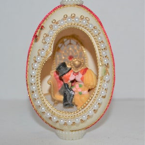 May include: A cream-coloured egg-shaped ornament, trimmed with red and gold-coloured detailing, and decorated with faux pearls. Inside, a miniature scene shows a couple in formal attire. The ornament has a decorative base. The egg is approximately 10 cm tall.