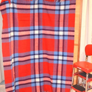 May include: A red plaid blanket with blue, white, and burgundy accents is hanging from a clothesline. The blanket has a fringed edge and is secured with clothespins. A red step stool is in the foreground.