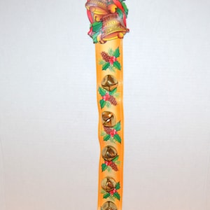 May include: A festive Christmas decoration with a series of golden bells on an orange vertical strip. The top features two bells, a red bow, and holly. The orange strip is decorated with holly, pinecones, and additional bells. Designed for hanging.