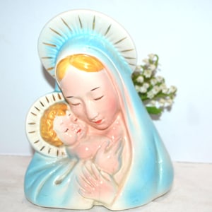 May include: A ceramic figurine of the Virgin Mary holding baby Jesus. The statue has a light blue robe, a white halo with gold detailing, and delicate facial features. The figurine is a religious item.