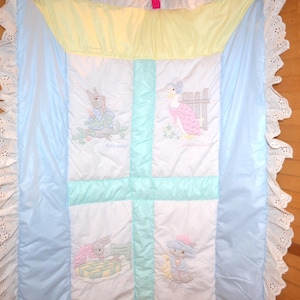 Könnte beinhalten: Eine gesteppte Babydecke mit einem weißen Mittelteil, der vier gestickte Szenen zeigt, umrandet von hellblauem Stoff und einem gelben oberen Bereich. Die Kanten sind rundherum mit einer weißen Lochstickerei-Rüsche versehen.