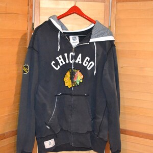 K&ouml;nnte beinhalten: Schwarzer Kapuzenpullover mit Rei&szlig;verschluss und dem Wort "CHICAGO" in wei&szlig;en Blockbuchstaben. Darunter befindet sich ein Logo eines indianischen H&auml;uptlings. Der Hoodie hat eine graue Kapuze und einen kleinen NHL-Patch auf dem linken &Auml;rmel.