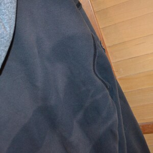 K&ouml;nnte beinhalten: Ein dunkelmarineblauer Kapuzenpullover mit grauer Kapuze. Der Kapuzenpullover hat ein schlichtes Design mit einer Nahtdetail. Der Hintergrund ist eine hellbraune Holzwand.