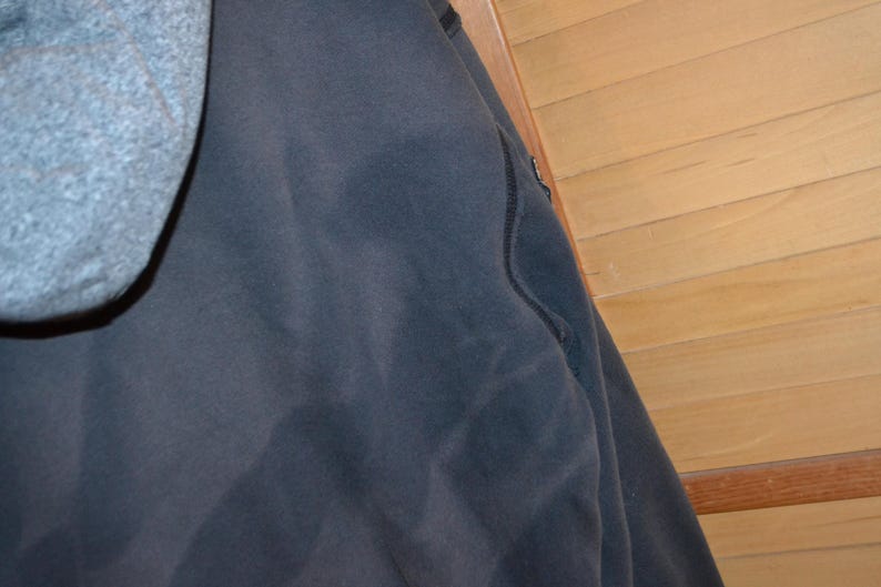 K&ouml;nnte beinhalten: Ein dunkelmarineblauer Kapuzenpullover mit grauer Kapuze. Der Kapuzenpullover hat ein schlichtes Design mit einer Nahtdetail. Der Hintergrund ist eine hellbraune Holzwand.