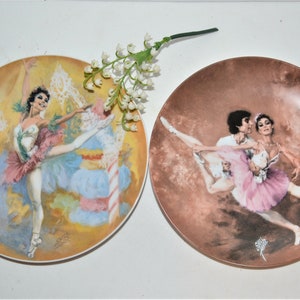 May include: Two porcelain plates with ballet dancer illustrations. The first plate features a solo ballerina in a pink and green tutu, dancing on a yellow and blue background. The second plate features a couple dancing in a pink and white tutu, on a brown background.
