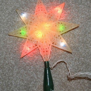 May include: A lighted Christmas tree topper in the shape of a star. The star is translucent with a textured surface and is lit with red, yellow, and green lights. It has a green base and a white electrical cord.