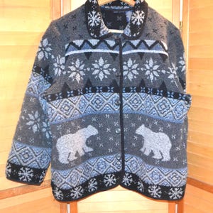 May include: Grey button-up cardigan with a winter theme. The cardigan features a pattern of snowflakes, geometric shapes, and polar bear silhouettes in shades of grey, black, and blue. The collar and cuffs are black.
