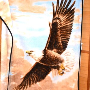 May include: A plush blanket featuring a soaring bald eagle with outstretched wings against a backdrop of clouds and a blue sky. The eagle is depicted in shades of brown, white, and tan, with a mountain range at the bottom. The blanket has a black border.