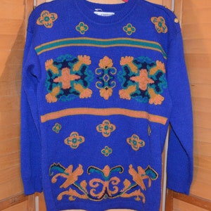 May include: A blue knitted sweater with a colourful embroidered design. The design features geometric shapes, flowers, and swirls in shades of orange, green, and brown.