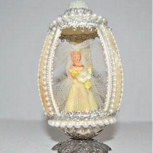 Op de afbeelding: Een eivormig decoratief ornament met een bruidsfiguur binnenin. Het ivoorkleurige ei is versierd met parels, zilveren randen en een sluier. De bruid draagt een gele jurk en houdt een boeket vast. Het ornament staat op een zilverkleurige basis.