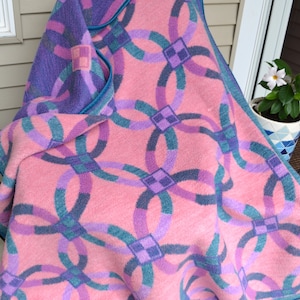 Puede incluir: Una manta tejida de color rosa, morado y turquesa con un patrón repetitivo de círculos entrelazados. La manta está doblada y drapeada sobre una silla.
