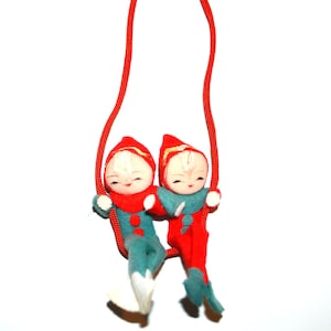 Puede incluir: Un adorno navideño vintage con dos muñecos de duendes. Los duendes visten trajes rojos y turquesas con sombreros a juego. Están suspendidos de un cordón rojo, creando una decoración navideña festiva y nostálgica.