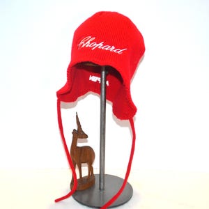 Könnte beinhalten: Eine leuchtend rote Strickmütze mit Ohrenklappen und langen Bändern. Das Wort "Chopard" ist in weißer Schrift auf die Vorderseite gestickt. Die Mütze wird auf einem Metallständer präsentiert. Eine kleine Holzfigur eines Hirsches befindet sich im Vordergrund.