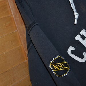 K&ouml;nnte beinhalten: Ein marineblauer Chicago Blackhawks Hoodie mit wei&szlig;en Buchstaben und einem Teamlogo. Der &Auml;rmel hat einen NHL-Patch. Der Hoodie hat einen Kordelzug und ein schwarz-wei&szlig; gestreiftes Kabel.