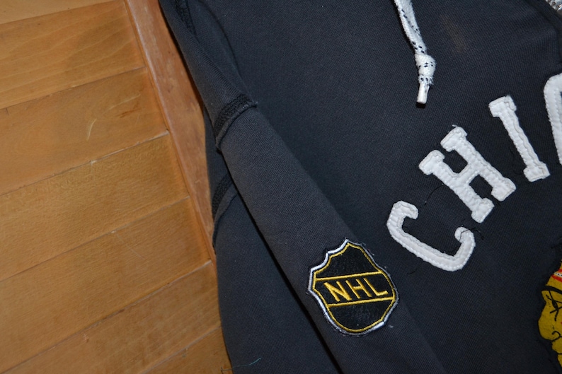 K&ouml;nnte beinhalten: Ein marineblauer Chicago Blackhawks Hoodie mit wei&szlig;en Buchstaben und einem Teamlogo. Der &Auml;rmel hat einen NHL-Patch. Der Hoodie hat einen Kordelzug und ein schwarz-wei&szlig; gestreiftes Kabel.