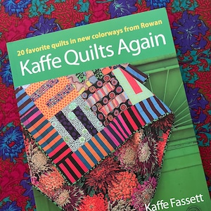 Może przedstawiać: Zielona książka zatytułowana "Kaffe Quilts Again" z kolorową kołdrą na wierzchu. Kołdra ma różne wzory i kolory, w tym pomarańczowy, różowy i czarny. Okładka książki zawiera również nazwisko autora, Kaffe Fassett.