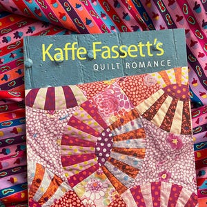 Puede incluir: Un libro titulado "Kaffe Fassett's Quilt Romance" con una cubierta turquesa y texto amarillo. El libro presenta un diseño de colcha de patchwork colorido en tonos de rosa, morado y naranja. El fondo es una tela vibrante con un patrón repetido de formas coloridas.