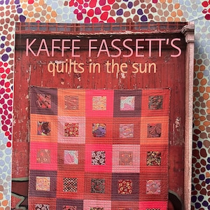 Op de afbeelding: Een boek getiteld "KAFFE FASSETT'S quilts in the sun" met een kleurrijk quiltontwerp op de omslag. De quilt heeft een patchwork-ontwerp met verschillende patronen en kleuren, waaronder rode, oranje en roze vierkanten. Het boek vermeldt ook "20 ontwerpen van Rowan voor patchwork en quilten."