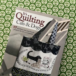 May include: A tote bag with a black and white floral pattern and polka dot handles rests on a book titled "It's Quilting Cats & Dogs." The bag features an embroidered design of a dachshund, sewing machine, and quilt.