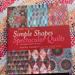 May include: A book titled "Kaffe Fassett's Simple Shapes Spectacular Quilts" with a red cover and colourful quilt designs. The book features 23 original quilt designs and photographs by Debbie Patterson. The title text is white and pink.