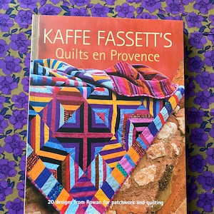 May include: A book titled "KAFFE FASSETT'S Quilts en Provence" with a vibrant quilt design on the cover. The quilt showcases geometric patterns in blue, orange, purple, and pink. The book rests on a purple floral patterned surface.