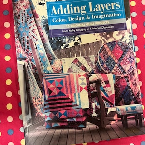May include: A book titled "Adding Layers: Color, Design & Imagination" featuring 15 original quilt projects by Kathy Doughty. The cover shows colourful quilts and a wooden chair. The background is a pink fabric with white, blue, and yellow polka dots.