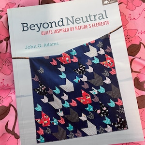 Op de afbeelding: Een boek getiteld "Beyond Neutral" met de ondertitel "Quilts Inspired by Nature's Elements" door John Q. Adams. De omslag toont een marineblauwe quilt met geometrische vormen in rood, wit, zwart en turquoise. De achtergrond is een roze bloemenstof.
