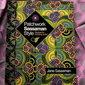 May include: A hardcover book titled "Patchwork Sassaman Style" with a colorful floral pattern. The cover features a black diamond with the title and author's name, Jane Sassaman. The book is on a pink and gray polka dot background.
