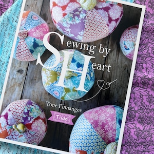 May include: A book titled Sewing by Heart by Tone Finnanger featuring a Tilda logo. The cover displays several round, fabric-covered pumpkin shapes made from various floral and patterned textiles in shades of pink, blue, green, and white.