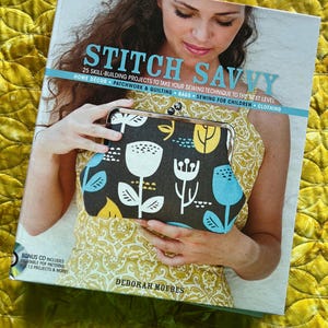 Könnte beinhalten: Eine Frau hält ein Exemplar von "Stitch Savvy" von Deborah Moebes, einem Buch über Nähprojekte. Das Buchcover zeigt den Titel in Türkis und eine Clutch mit schwarzen und weißen Blumen. Der Hintergrund ist ein gelber, gesteppter Stoff.