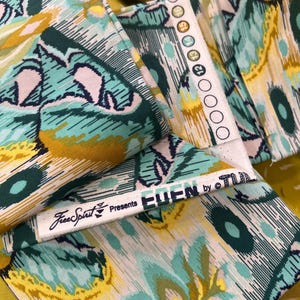 Puede incluir: Una pieza de tela doblada con un patrón floral abstracto en tonos verde azulado, amarillo mostaza y blanco. El orillo muestra el texto Free Spirit Presents EDEN by TULA PINK y una serie de círculos numerados del 12 al 16 en tinta negra.