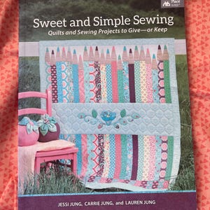 May include: A softcover book titled Sweet and Simple Sewing: Quilts and Sewing Projects to Give—or Keep by Jessi Jung, Carrie Jung, and Lauren Jung. The cover features a quilt with a pencil pattern, a pink chair, and two fabric pumpkins.