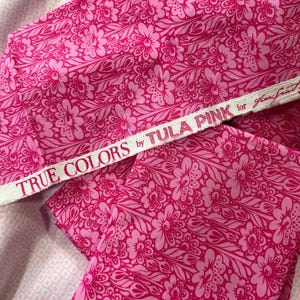 May include: Bright pink floral fabric with a detailed pattern. The fabric features a repeating design of flowers and leaves in varying shades of pink. A white ribbon with the text "TRUE COLORS by TULA PINK" is visible.
