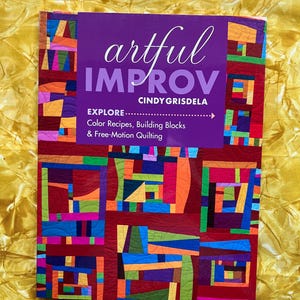 Könnte beinhalten: Ein Buch mit dem Titel "artful IMPROV" von Cindy Grisdela, das ein farbenfrohes Quilt-Design zeigt. Das Cover ist lila mit weißem Text, und das Quilt-Design enthält rote, orangefarbene, blaue, grüne und gelbe geometrische Formen. Das Buch befasst sich mit Farbrezepten, Bausteinen und Freihandquilten.