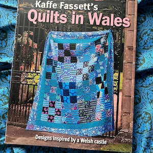 May include: A softcover book titled Kaffe Fassett's Quilts in Wales, Designs inspired by a Welsh castle. The cover features a patchwork quilt in shades of blue, teal, and black, displayed against a background of a black metal gate and stone pillar.