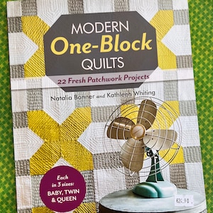 Op de afbeelding: Een boek getiteld "MODERN One-Block QUILTS" met een patchwork-ontwerp in geel, grijs en wit. De boekomslag bevat tekst en wordt vergezeld door een vintage ventilator met een groene basis en messing bladen. Het boek is verkrijgbaar in drie maten: BABY, TWIN & QUEEN.
