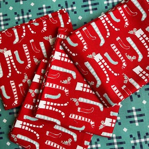 May include: Three folded pieces of red fabric featuring a repeating pattern of white and light blue striped stockings, solid light blue stockings, and small sprigs of green holly with red berries, layered over a teal background with a white grid.